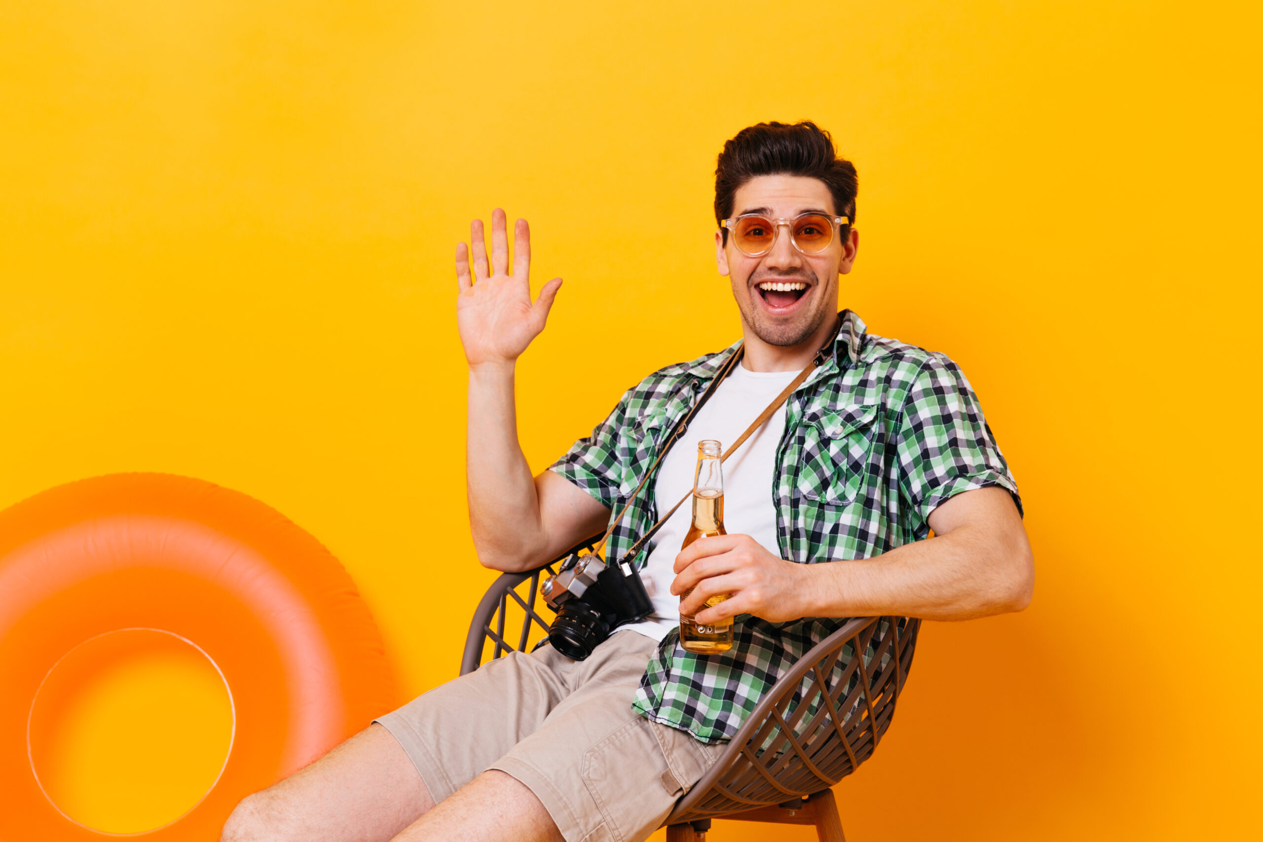 Charming man in green plaid shirt waving his hand, laughing, holding bottle of beer and retro camera on orange background. Charming man in green plaid shirt waving his hand, laughing, holding bottle of beer and retro camera on orange background